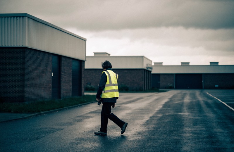 Iemand in een geel veiligheidshesje loopt over een bedrijventerrein langs gesloten roldeuren.