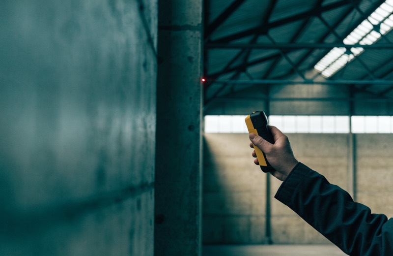 Een laserafstandsmeter in de hand van de taxateur, gericht op de binnenwand van een lege bedrijfshal met de rode meetstip op de wand aan de overkant.