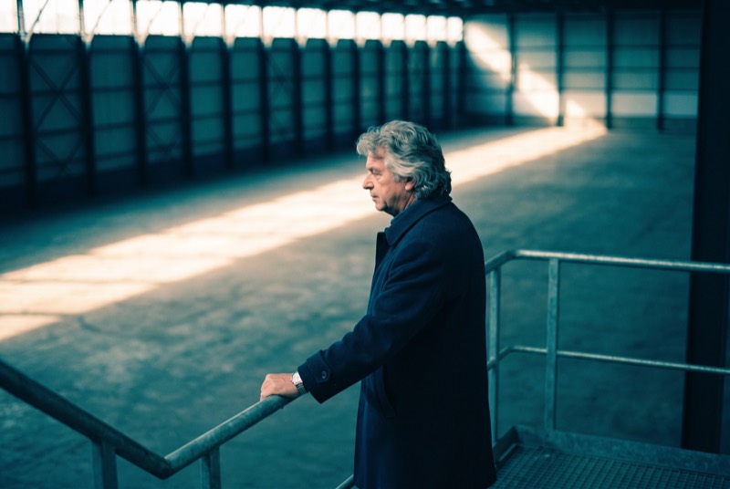 Een taxateur staat op een stalen bordes boven aan een industrietrap met een hand op de buisleuning en kijkt uit over de lege betonvloer beneden hem.