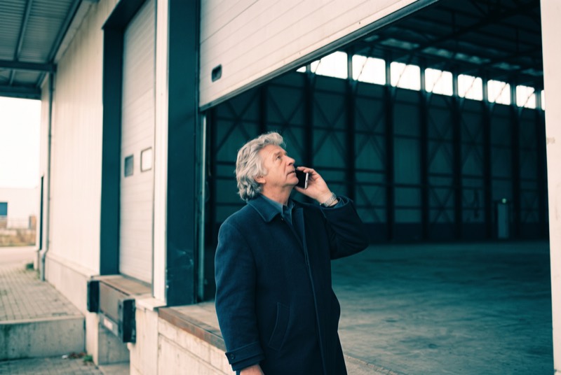 Een taxateur staat op een betonnen laadperron naast de open roldeur van een lege loods en spreekt in zijn telefoon terwijl hij langs de rand van de dagopening omhoog kijkt.