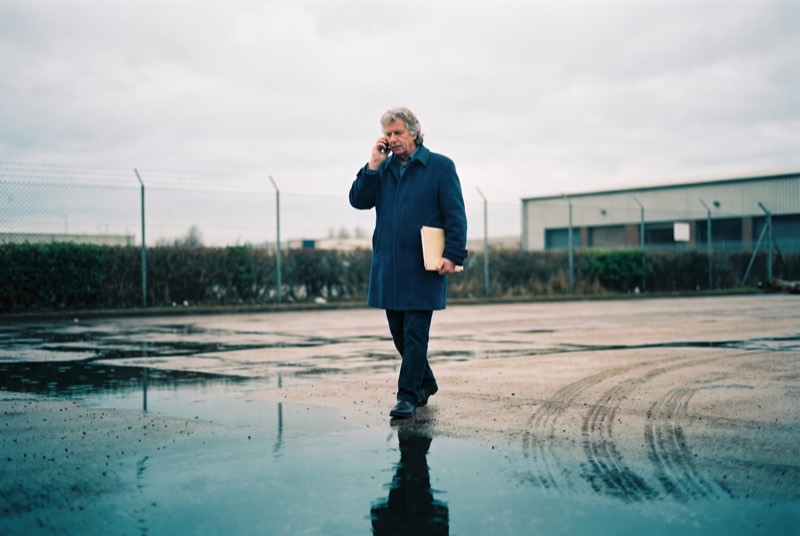Een taxateur loopt over een breed leeg asfaltterrein achter een bedrijfsunit met een map onder zijn arm; het asfalt is nat en weerspiegelt de grijze lucht.