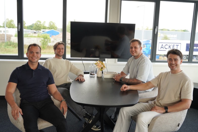 Vier collega's zitten rond een ovale tafel bij een raamwand met uitzicht op het bedrijventerrein in Oss.